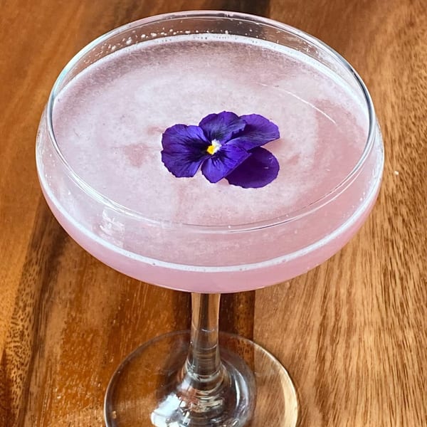 Pink cocktail in a coupe glass, garnished with a vibrant purple edible flower on a wooden table.